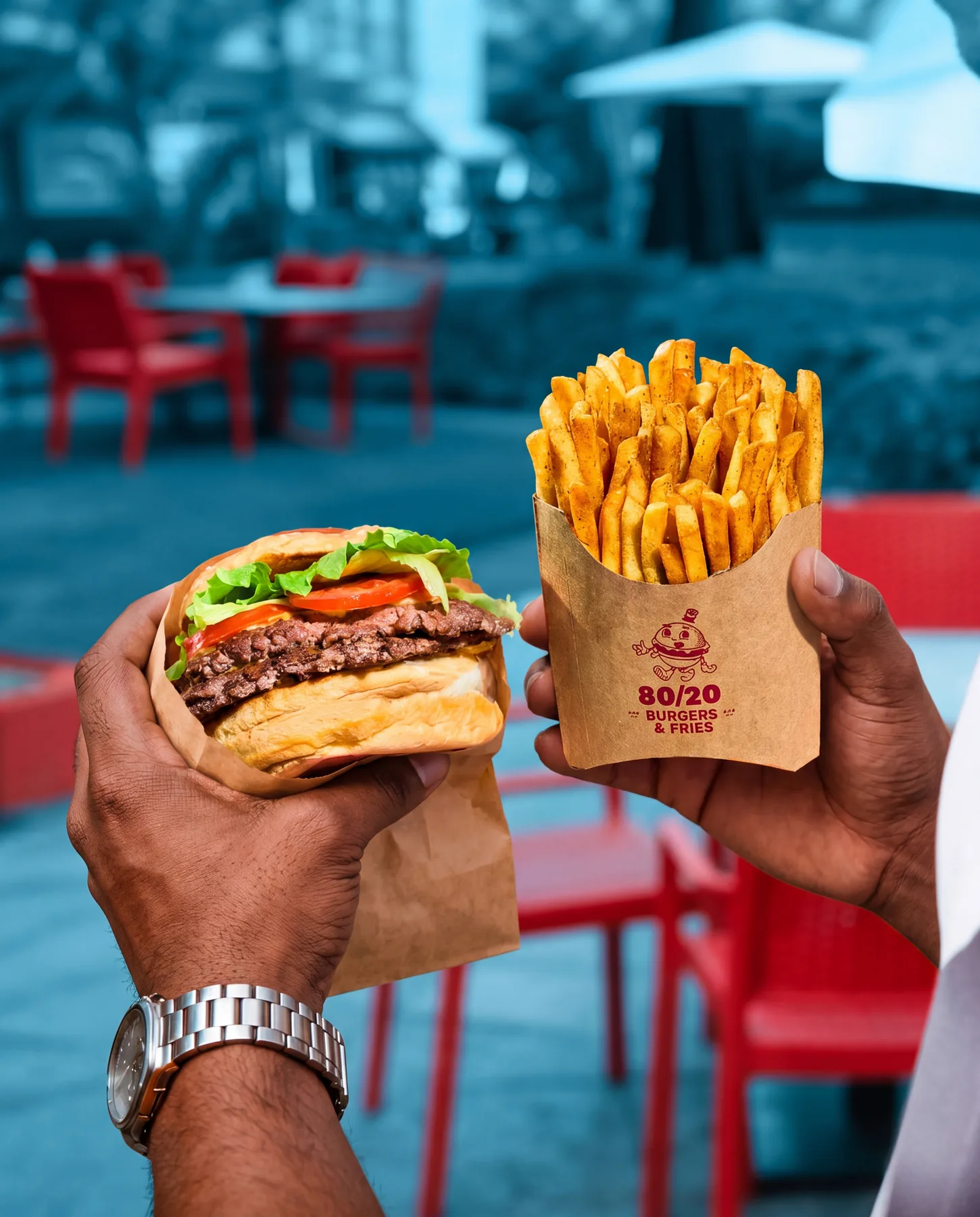 Two hands holding up a burger and fries at a red outdoor table